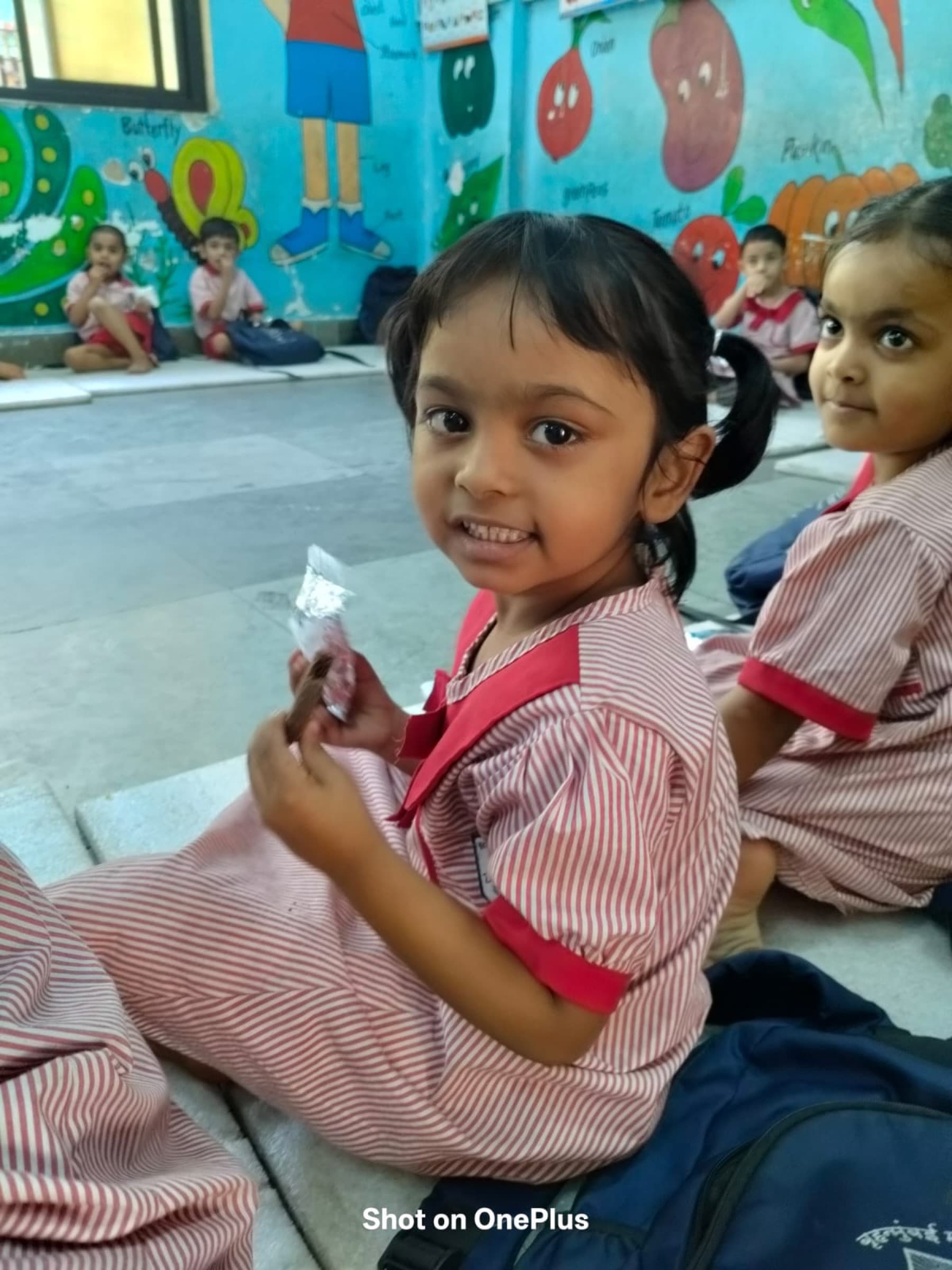A girl in her striped anganwadi uniform smiles while holding a piece of the protein snack.