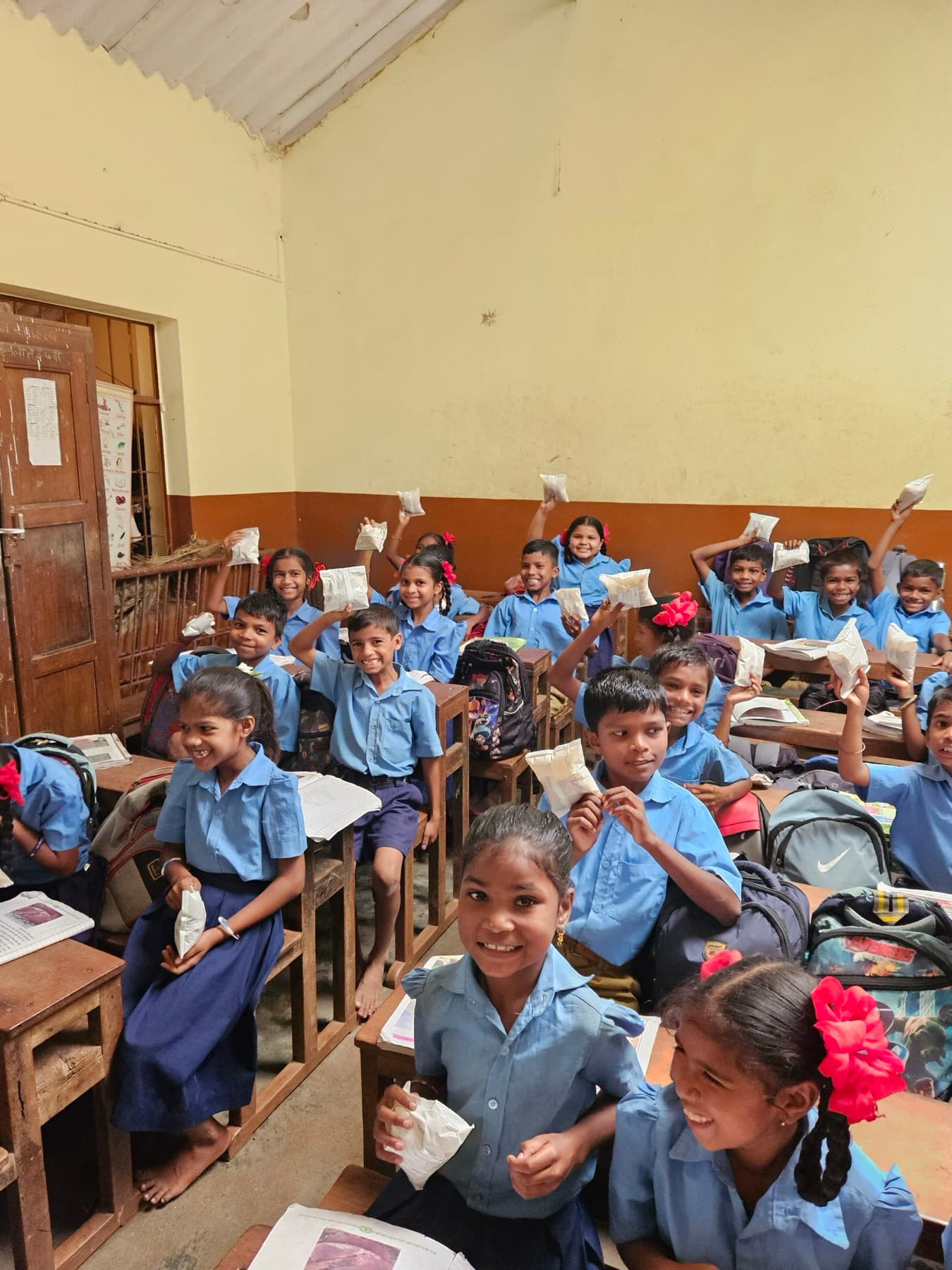 Children in blue uniforms hold up their nutrition packets in a village classroom.