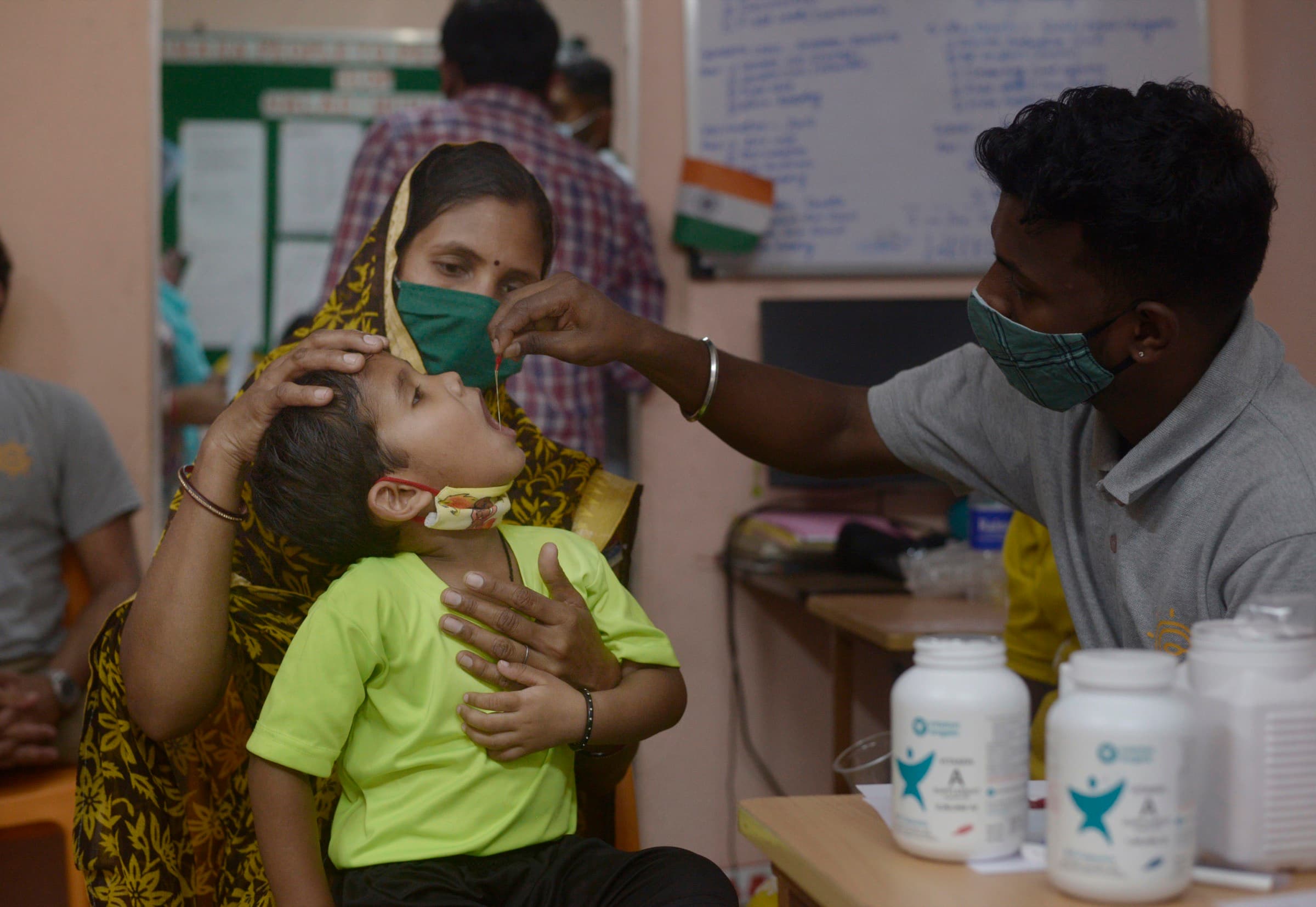 A mother steadies her child while a masked health worker gives a Vitamin A drop, part of a routine screening visit.