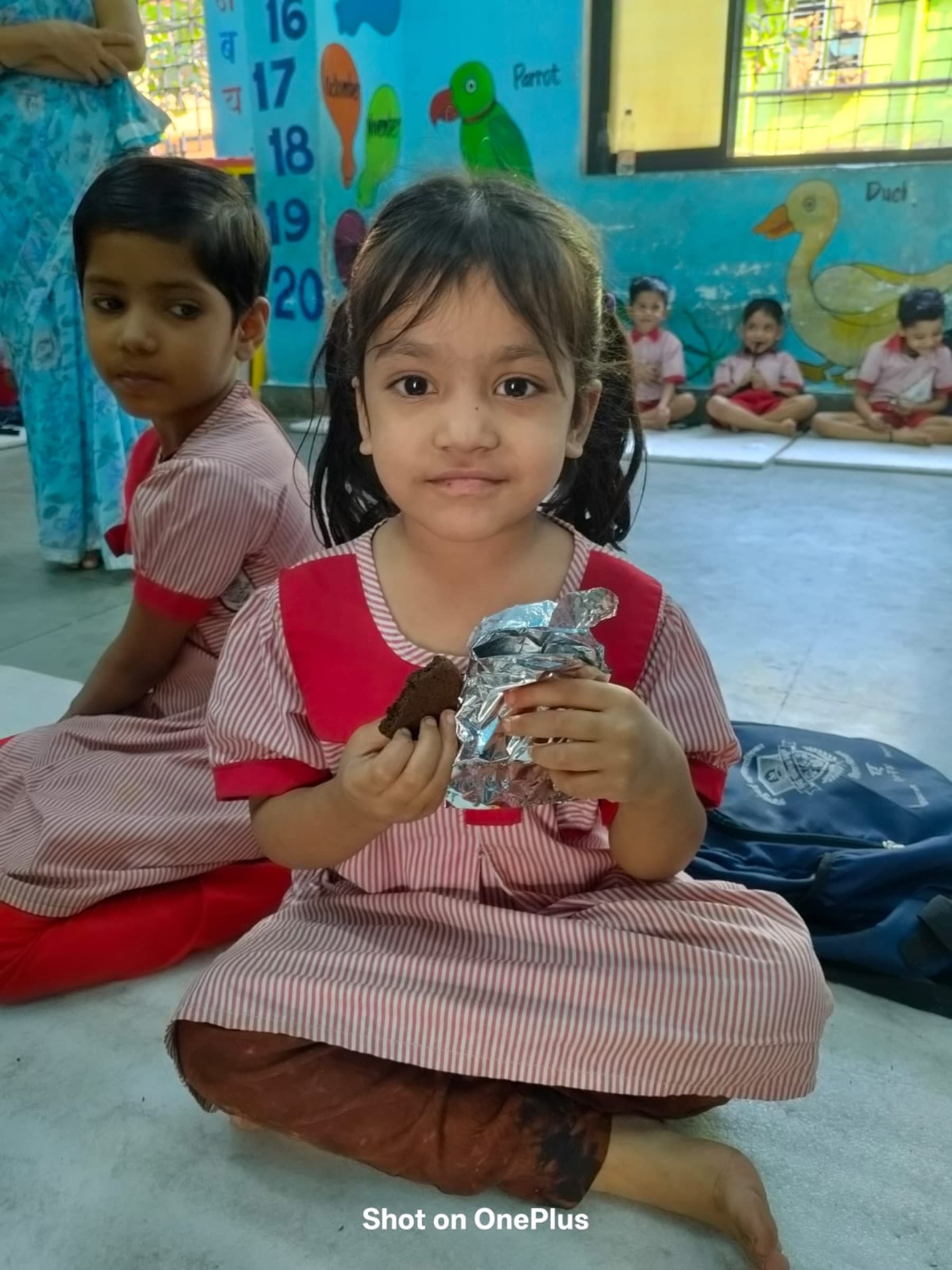 A young girl in her striped anganwadi uniform sits with classmates on the floor, holding a piece of the protein snack during a session.
